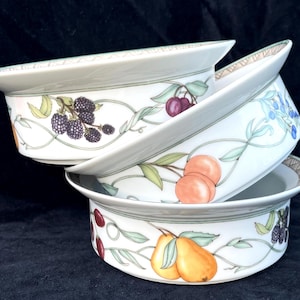 May include: A stack of three white ceramic bowls featuring a decorative fruit pattern. The design includes illustrations of blackberries, cherries, peaches, and pears connected by green vines. Each bowl has a wide rim with a geometric border.