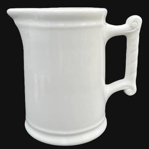 May include: White ceramic pitcher with a handle that has a decorative scroll design. The pitcher has a rounded body and a wide mouth.