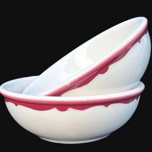 May include: Two stacked white ceramic bowls featuring a decorative pink scalloped rim design. The bowls have a smooth, glossy finish and a rounded shape, with the top bowl angled to reveal the interior and the bottom bowl resting on a base.