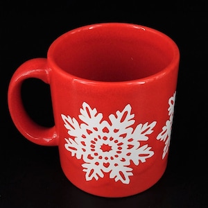 May include: A red ceramic mug with a white snowflake design. The mug has a handle and is perfect for hot beverages.
