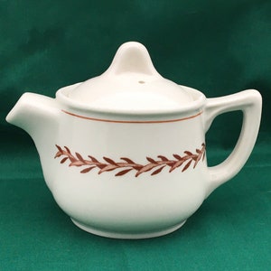 May include: Cream-colored ceramic teapot with a brown leaf pattern around the body. The teapot has a spout, handle, and a lid with a small knob. A brown line accents the rim.