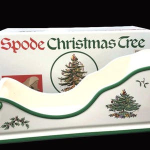 May include: A white ceramic sleigh with green trim and Christmas tree illustrations. The sleigh is in front of a box that reads "Spode Christmas Tree" in red and green lettering. Festive holiday decor.