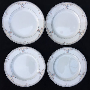 May include: Four white ceramic plates with a delicate floral and gold rim design. Each plate features a central white area surrounded by a patterned border with small flowers in shades of pink, blue, and yellow. The plates are arranged in a square formation.