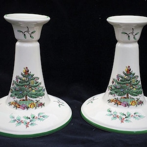May include: Two white ceramic Christmas candle holders. Each holder features a green Christmas tree design with ornaments and presents. The base and rim are trimmed in green, with holly and mistletoe accents.