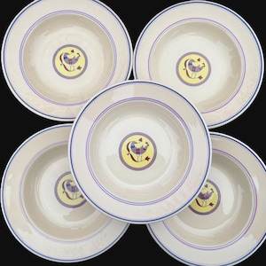 May include: Five cream-colored ceramic plates with a blue and purple rim design. Each plate features a central emblem of a bird in purple, yellow, and red. The plates are arranged on a black background.