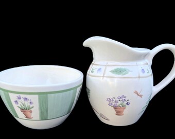 May include: A white ceramic pitcher and bowl set with a garden theme. The pitcher has a handle and spout, decorated with floral and dragonfly motifs. The bowl has a green striped design with potted flowers. The set is ideal for serving.