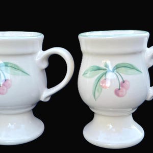 May include: Two white ceramic mugs with a pedestal base and curved handles. Each mug features a hand-painted design of cherries and green leaves. The rims are trimmed with a light blue accent color. The mugs are set against a black background.