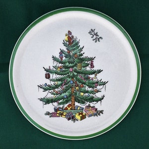 May include: A white ceramic plate with a green rim features a hand-painted Christmas tree with ornaments, presents, and a small figure at the top. The tree is surrounded by holly leaves and berries.