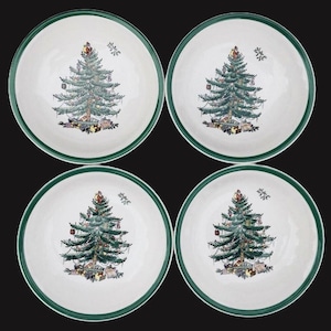 May include: Four white ceramic plates with a green rim. Each plate features a detailed illustration of a decorated Christmas tree with presents underneath. The plates are arranged on a black background.