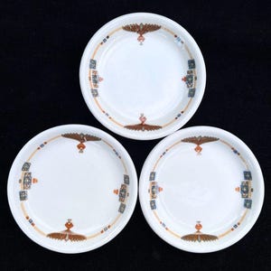May include: Three white ceramic saucers with a brown, orange, and blue geometric design. The saucers are empty and have a slightly raised rim.