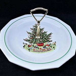 May include: White, octagonal serving platter with a green rim and a gold handle. The platter features a Christmas tree design with a village scene at the base. The handle is gold-colored.
