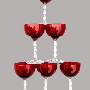 May include: A pyramid of six red-colored champagne glasses. The glasses have a rounded bowl shape and sit atop clear, twisted stems. The glasses are arranged in a tiered formation, with one glass at the top, two in the middle, and three at the bottom.