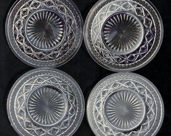 May include: Set of four clear glass dessert plates with a decorative, geometric pattern. The plates have a raised, starburst design in the center.