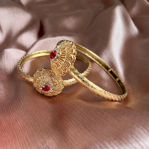 May include: Two gold-toned bangles featuring intricate filigree metalwork and a central floral-shaped ornament. Each ornament is set with a single round red gemstone surrounded by small clear stones, with a textured pattern along the band.