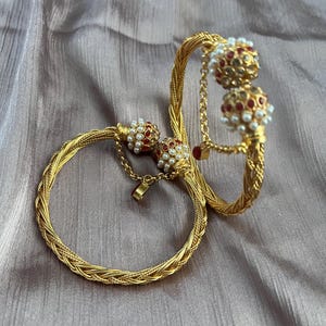 Peut inclure: Deux bracelets dorés avec un design tressé complexe. L'un est fermé, l'autre ouvert et relié par une chaîne. Les deux présentent des ornements perlés rouges et blancs, créant un ensemble de bijoux élégant.
