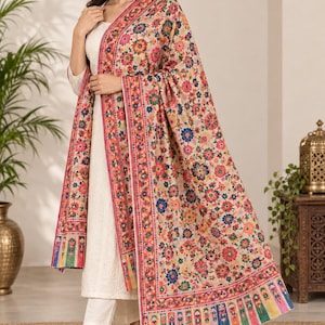 Puede incluir: Un conjunto de túnica de manga larga y pantalones de color crema, combinado con un gran chal rectangular con un vibrante estampado floral en tonos rojos, naranjas, azules y verdes, con un borde decorativo y un dobladillo con flecos.