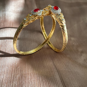 Peut inclure: Deux bracelets dorés avec des motifs de filigrane complexes. Chaque bracelet est orné d'une pierre rouge centrale entourée de petites pierres transparentes. Les bracelets reposent sur une surface texturée brun clair.