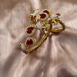 May include: Ornate gold-coloured bracelet with floral and leaf detailing. It features teardrop-shaped, deep red gemstones and small, white pearls. The design includes a chain and is set against a peach-coloured fabric.
