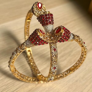 Può includere: Due braccialetti color oro con dettagli intricati e gemme rosse e chiare. I braccialetti sono adornati da un motivo floreale e sono posti su uno sfondo marrone chiaro. Le gemme sono raggruppate per creare un effetto scintillante.