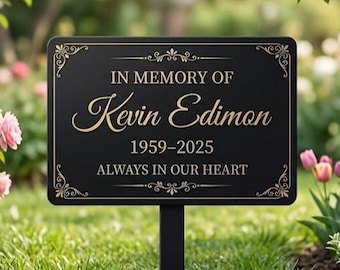 Puede incluir: Una placa conmemorativa rectangular negra sobre estaca con texto en color dorado que dice IN MEMORY OF Kevin Edimon 1959-2025 ALWAYS IN OUR HEART, con esquinas decorativas doradas y un borde fino, montada sobre un poste metálico negro.
