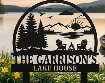Puede incluir: Un letrero de metal negro con una parte superior semicircular que presenta un paisaje cortado con láser de pinos, montañas, un sol, dos sillas, una fogata y pájaros. El texto dice THE GARRISON'S LAKE HOUSE. El letrero tiene dos estacas.