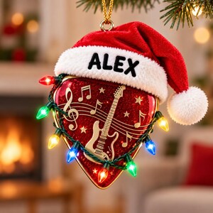 Könnte beinhalten: Ein herzförmiger Weihnachtsbaumschmuck mit roter Glitzeroptik, einer goldenen E-Gitarre und Notenmotiven. Der Anhänger ist mit einer roten Weihnachtsmütze mit dem Namen ALEX versehen und von einer Lichterkette mit bunten Lichtern umwickelt.