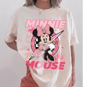 May include: Cream-colored t-shirt featuring a graphic of Minnie Mouse in a pink and black design. The text "MINNIE MOUSE" is displayed in pink. The shirt is paired with red and white checkered shorts.