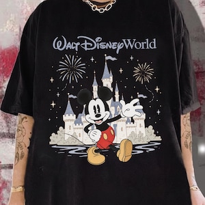 May include: Black t-shirt featuring Mickey Mouse waving in front of Cinderella's castle with fireworks and the text "Walt Disney World". The design is in a cartoon style with a blue, white, red, and yellow color palette.