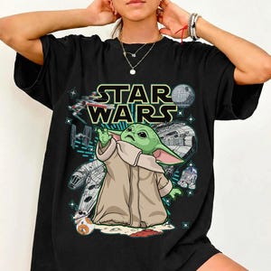 Op de afbeelding: Zwart T-shirt met een Star Wars-afbeelding. Het ontwerp bevat de tekst "STAR WARS" boven een cartoonafbeelding van Grogu, ook bekend als Baby Yoda, met verschillende Star Wars-elementen.