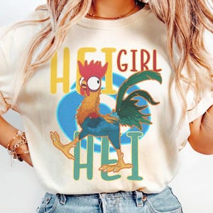 May include: Cream-colored t-shirt featuring a cartoon rooster with the text "HEI GIRL HEI". The rooster is in motion, with a red comb, yellow and orange feathers, and blue accents. The text is in yellow and teal.
