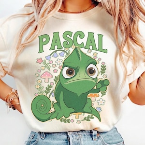 Könnte beinhalten: Cremefarbenes T-Shirt mit einem grünen Cartoon-Chamäleon und dem Namen "PASCAL" darüber in grünen Buchstaben. Das Chamäleon ist von bunten Blumen und Pilzen umgeben. Ein lässiges Kleidungsstück.