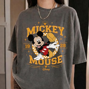 May include: A gray t-shirt featuring a graphic of Mickey Mouse in a dynamic pose. The design includes the words "Mickey Mouse" in yellow, along with the year "1928" and the Disney logo. The shirt has short sleeves.