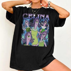 Puede incluir: Camiseta negra con un gráfico de dibujos animados. El gráfico presenta el nombre "CELINA" en morado y varias figuras alienígenas en verde y morado. Las figuras tienen un ojo y visten vestidos y tops. El fondo tiene un diseño azul y morado.