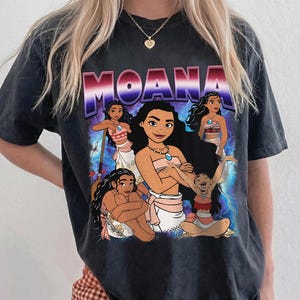 Puede incluir: Camiseta gris oscuro con la palabra "MOANA" en una fuente degradada púrpura y roja. La camiseta muestra un gráfico del personaje de Moana y otros personajes de la película, con un fondo de rayos azules y blancos.