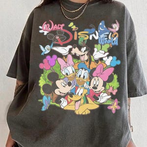 May include: Dark gray t-shirt featuring a colorful graphic of Disney characters, including Mickey Mouse, Minnie Mouse, Donald Duck, and Goofy. The text "Walt Disney World" is also displayed in a playful font. The shirt has short sleeves and a relaxed fit.