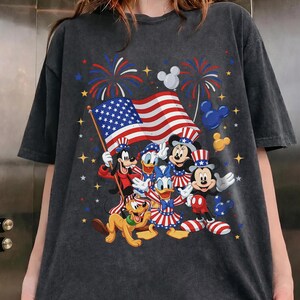 May include: Dark gray t-shirt featuring a patriotic design with Disney characters. The graphic includes Mickey Mouse, Minnie Mouse, Donald Duck, Goofy, and Pluto, along with an American flag and fireworks.