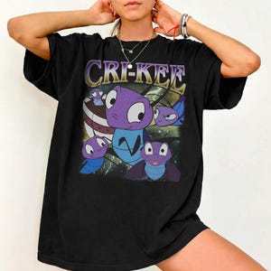 Może przedstawiać: Czarny t-shirt z grafiką postaci Cri-Kee z Mulan. Projekt zawiera imię postaci w fioletowej i złotej czcionce, a także obrazy postaci w różnych pozach. Koszulka ma swobodny, obszerny krój.