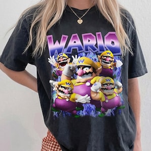 Puede incluir: Camiseta negra con un gráfico del personaje Wario de la serie de videojuegos. El diseño incluye múltiples figuras de Wario con sombreros amarillos y monos morados, con el nombre "WARIO" en letras moradas grandes.