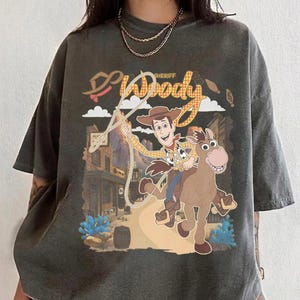 May include: Dark gray t-shirt featuring a graphic of Sheriff Woody from Toy Story riding a horse, with the word "Woody" and "Sheriff" in a western-style font. The design includes a rope and a cityscape background.