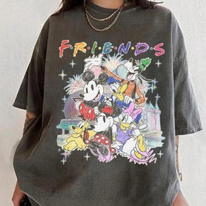 May include: Dark gray t-shirt featuring the word "FRIENDS" in colorful lettering above a graphic of Disney characters including Mickey Mouse, Minnie Mouse, Donald Duck, and Goofy. The design includes fireworks and a castle.