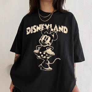 May include: Black t-shirt featuring a distressed graphic of Minnie Mouse and the word "Disneyland" in a vintage font. The design is in a cream color. The shirt has short sleeves and a relaxed fit.