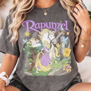 Könnte beinhalten: Dunkelgraues T-Shirt mit einer farbenfrohen Grafik von Rapunzel und anderen Charakteren aus dem Film. Das Wort "Rapunzel" ist lila über der Grafik aufgedruckt. Das T-Shirt hat kurze Ärmel und einen Rundhalsausschnitt.