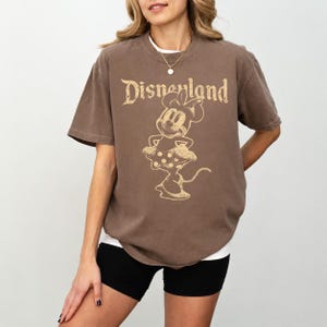 K&ouml;nnte beinhalten: Braunes T-Shirt mit dem Wort "Disneyland" in einer Vintage-Schriftart und einer Minnie-Maus-Grafik. Das Shirt hat kurze &Auml;rmel und eine lockere Passform. Das Model tr&auml;gt schwarze Shorts.