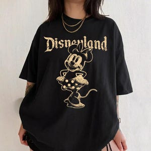 K&ouml;nnte beinhalten: Schwarzes T-Shirt mit einem goldenen Minnie Mouse-Motiv und dem Wort "Disneyland" in einer Vintage-Schriftart. Das T-Shirt hat kurze &Auml;rmel und eine lockere Passform.