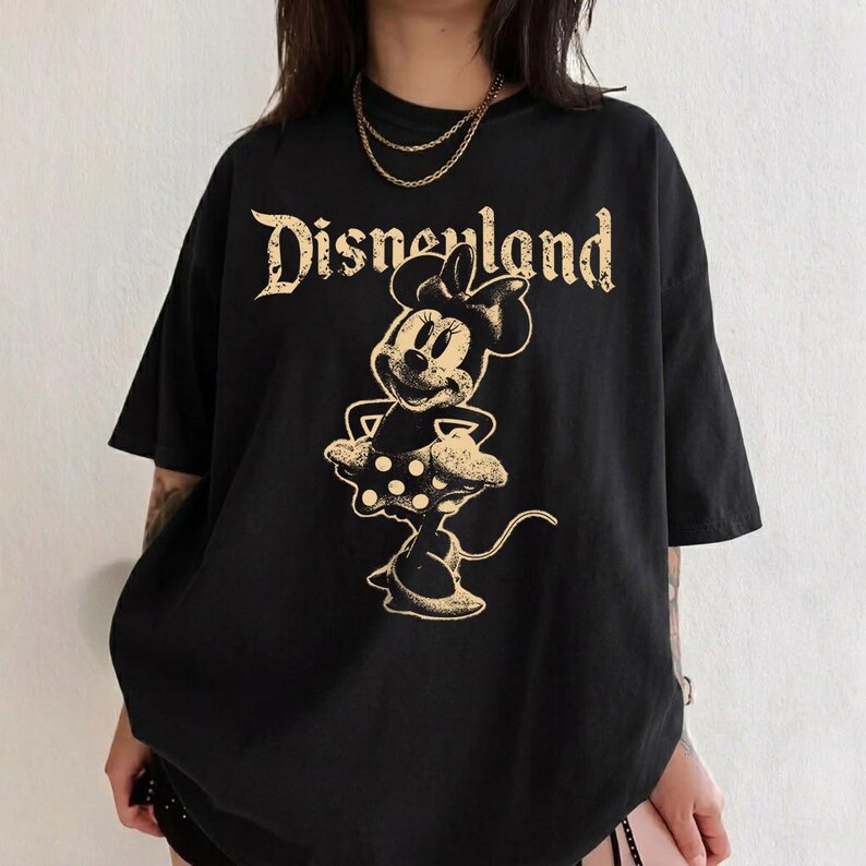 K&ouml;nnte beinhalten: Schwarzes T-Shirt mit einem goldenen Minnie Mouse-Motiv und dem Wort "Disneyland" in einer Vintage-Schriftart. Das T-Shirt hat kurze &Auml;rmel und eine lockere Passform.