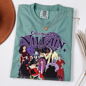 K&ouml;nnte beinhalten: Ein salbeigr&uuml;nes T-Shirt mit einem Grafikdruck verschiedener animierter B&ouml;sewichte. Der Text Chillin Like A Villain steht &uuml;ber den Figuren. Das T-Shirt hat ein Comfort Colors Markenetikett am Hals und einen Rundhalsausschnitt mit violetten Akzenten.