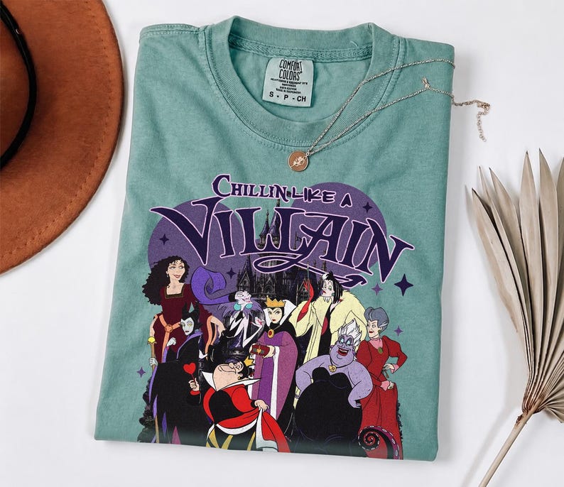 K&ouml;nnte beinhalten: Ein salbeigr&uuml;nes T-Shirt mit einem Grafikdruck verschiedener animierter B&ouml;sewichte. Der Text Chillin Like A Villain steht &uuml;ber den Figuren. Das T-Shirt hat ein Comfort Colors Markenetikett am Hals und einen Rundhalsausschnitt mit violetten Akzenten.