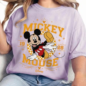May include: A lilac-colored t-shirt featuring a graphic of Mickey Mouse. The design includes the words "Mickey Mouse" in yellow, with the year "1928" and the Disney logo. Mickey is depicted in his classic pose, with red shorts and white gloves.