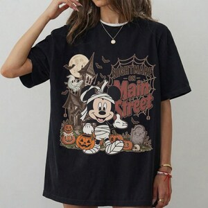 May include: Black t-shirt featuring a Halloween-themed graphic. The design includes Mickey Mouse dressed as a mummy, pumpkins, a haunted house, and the text "Nightmare on Main Street". The shirt has a vintage, distressed look.