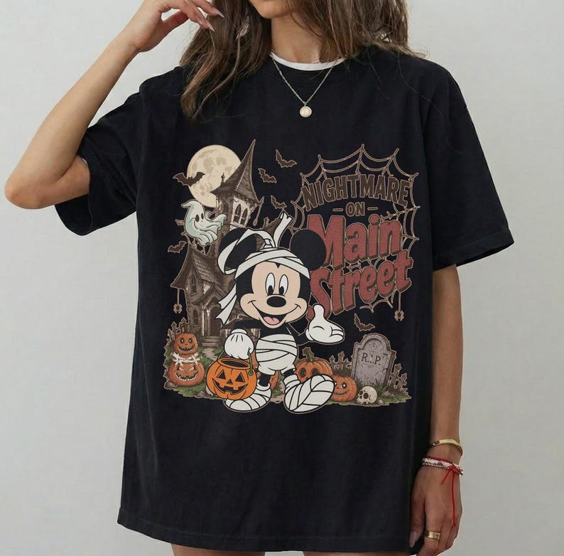 May include: Black t-shirt featuring a Halloween-themed graphic. The design includes Mickey Mouse dressed as a mummy, pumpkins, a haunted house, and the text "Nightmare on Main Street". The shirt has a vintage, distressed look.